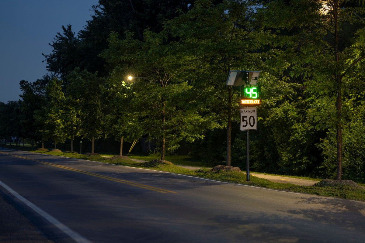 Afficheur de vitesse radar solaire indiquant 45 km/h devant un panneau de limite de 50 km/h, en bordure d’une route boisée.